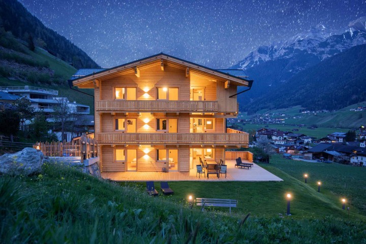 Illuminated wooden house in the valley, surrounded by lights on the lawn, under a twinkling starry sky at night.