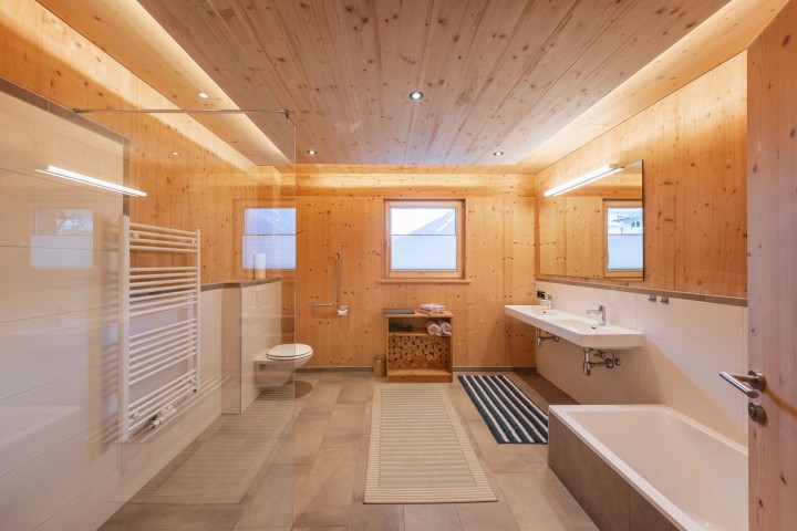 Mit Holz verkleidetes Badezimmer mit Glasdusche, Badewanne, WC, zwei Waschbecken und Fenster